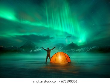 Ein Mann, der in den wilden nördlichen Bergen mit einem beleuchteten Zelt campt und eine spektakuläre grüne Nordlicht-Aurora-Anzeige betrachtet. Fotomontage.