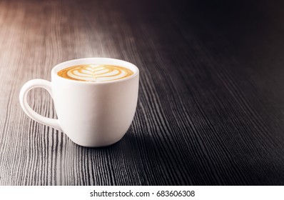 Tutup cangkir kopi putih dengan busa seni latte berbentuk hati di atas meja kayu hitam dekat jendela dengan bayangan terang di atas meja di kafe.