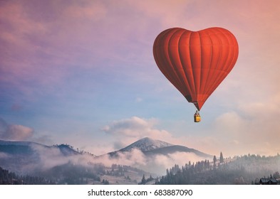 Schöne rote Luftballon-Herzform gegen blauen und rosafarbenen Pastellhimmel an einem sonnigen, hellen Morgen. Nebelige Berge im Hintergrund. Romantische Reise am Valentinstag. Thema Sport- und Erholungsreisen