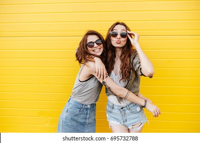 Bild von zwei jungen glücklichen Freundinnen, die über einer gelben Wand stehen. Blick in die Kamera, die Küsse bläst.