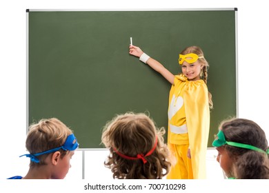nxënës të lumtur me kostume superheroish që studiojnë pranë dërrasës së zezë pas