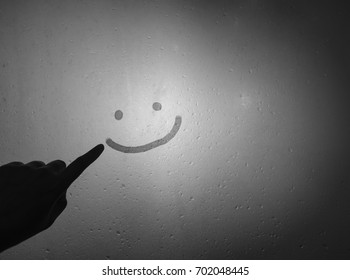 Mano de mujer escribir cara sonriente y feliz en el cristal de la ventana con lluvia o gotas de agua y vapor, en blanco y negro, humor oscuro