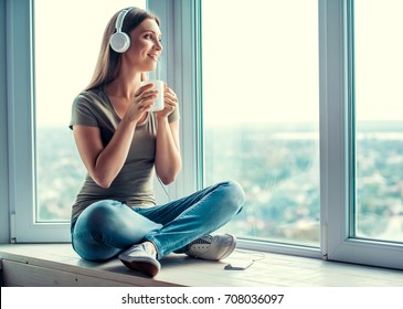 Beautiful woman in casual clothes and headphones is listening to music, holding a cup and smiling while sitting on window sill at home
