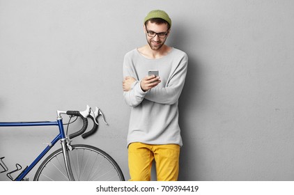 Vrolijke jonge blanke hipster met stoppels gekleed stijlvol spelend videogame onweb-enabled elektronisch gadget. Gelukkig lachende man fietser in glazen surfen op internet via wifi op mobiele telefoon
