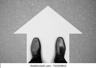 zapatos negros de pie sobre el piso de concreto asfáltico y señal de dirección blanca para seguir adelante.