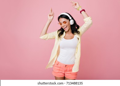 Inspired slim brunette girl in sunglasses funny dancing on pink background and waving hands. Laughing dark-haired young woman in yellow shirt enjoying music in headphones with eyes closed.