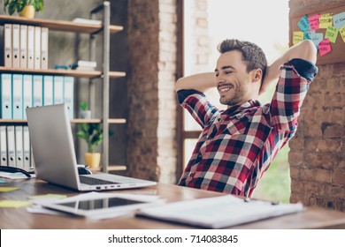 Chàng trai trẻ tóc nâu vui vẻ đang xem màn hình máy tính xách tay của anh ấy, tại nơi làm việc của anh ấy, với hai cánh tay sau đầu, nghỉ ngơi, mỉm cười, trong văn phòng
