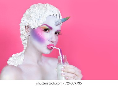 ピンクの背景の虹のユニコーンは、ボトルから牛乳を飲みます。若い女性のクローズ アップの肖像画。ハロウィンの衣装