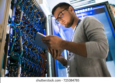 Retrato de ángulo bajo de joven usando tableta digital de pie junto al gabinete del servidor mientras trabaja con supercomputadora en luz azul