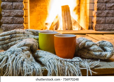 Dua cangkir untuk teh atau kopi, barang-barang wol di dekat perapian yang nyaman, di rumah pedesaan, liburan musim dingin, horizontal.
