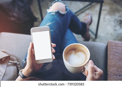 Mockup image of woman's hands holding white mobile phone with blank screen on thigh and coffee cup in cafe