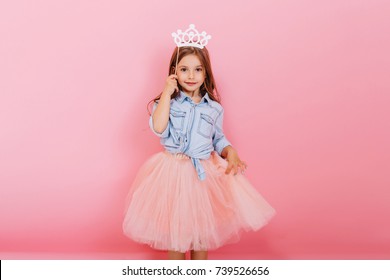 Niña alegre con cabello largo morena en falda de tul con corona de princesa en la cabeza aislada sobre fondo rosa. Celebrando el carnaval brillante para niños, fiesta de cumpleaños, divertirse con un niño lindo