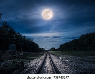 Railroad tracks through the woods at night. Beautiful blue sky and full moon above silhouettes of trees and railway. Serenity nature background. Outdoor at nighttime. The moon taken with my own camera