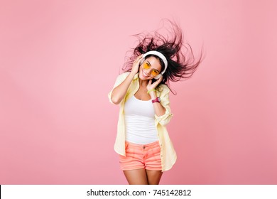 Glad latin girl in colorful clothes posing with black hair waving and laughing. Pleased asian female model in headphones having fun in summer studio photoshoot.