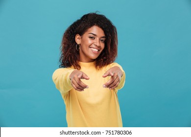 Photo of happy young african lady dressed in warm sweater standing isolated over blue background. Looking camera while pointing to you.