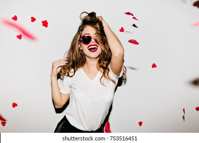 Indoor portrait of amazing caucasian female model in trendy t-shirt touching her long shiny hair. Laughing refined woman in sunglasses having fun with red confetti.