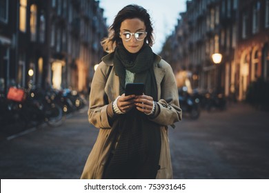 Hermosa joven elegante caminando por las calles de la ciudad de noche usando un moderno smartpone, una bloguera hipster escribiendo un mensaje de texto en su celular