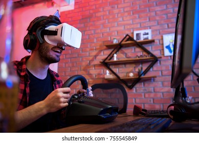 Computer Simulatioun. Gléckleche jonke bäertege Gamer a virtueller Realitéit Brille dréckt Opreegung aus wärend hien Autosportvideospill mam Lenk genéisst