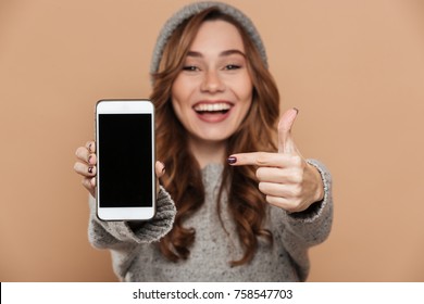 Portrait of cute brunette girl in warm hat and sweater pointing with finger on blank mobile screen, selective focus