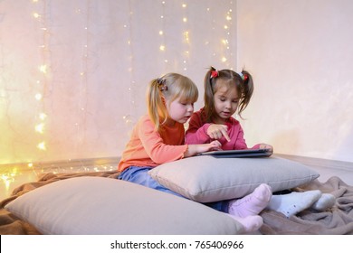 Cute little girls play in computer games on device or watch cartoons, press something on touch screen of tablet, smile and communicate with each other, sitting on rug in bright room with flickering