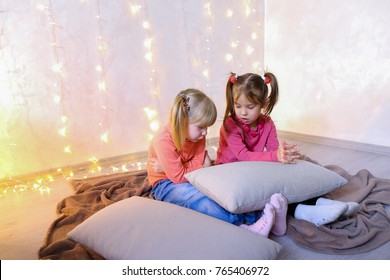 Cute little girls play in computer games on device or watch cartoons, press something on touch screen of tablet, smile and communicate with each other, sitting on rug in bright room with flickering