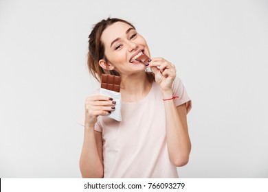 Portret van een tevreden mooi meisje dat chocoladereep bijt en naar camera kijkt die op witte achtergrond wordt geïsoleerd