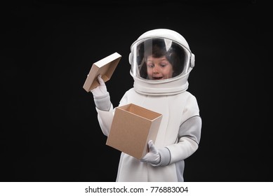 Best surprise. Portrait of little cosmonaut wearing helmet and protective costume is standing and opening cardboard box while feeling wonder and curiosity. Isolated background