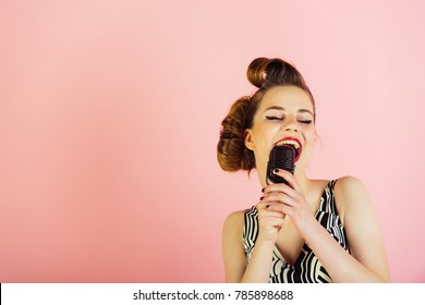Gruaja këngëtare me flokë dhe grim elegant retro. Vajza me syze këndon në mikrofon. Bukuria dhe moda vintage. Ngjiz vajzën e re në sfond rozë, radio. Muzikë, look dhe stil retro, pinup.