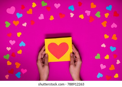 Las manos de la mujer sostienen una tarjeta de felicitación con un corazón de fondo rosa con corazones de papel multicolores alrededor. Regalos de vacaciones