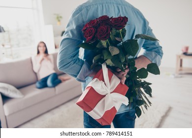 Unexpected moment in routine everyday life! Cropped photo of man's hands hiding holding chic bouquet of red roses and gift with white ribbon behind back, happy woman is on blurred background