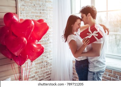 Beautiful young couple at home. Hugging, kissing and enjoying spending time together while celebrating Saint Valentine's Day with gift box in hand and air balloons in shape of heart on the background.