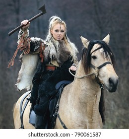 Schöne wütende skandinavische Wikinger-Kriegerin mit Kriegsmake-up in traditioneller Kleidung, die ein Pferd in einem Wald mit Axt in der Hand reitet und droht. Lagertha Blick. Serie