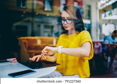 腕時計のディスプレイを見て、ラップトップでメールをチェックしながら時間を管理する眼鏡をかけた若いビジネスウーマン。締め切り前にネットブックで記事に取り組んでいる手元のスマートウォッチを持つフリーランサー
