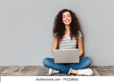 Retrato de una mujer satisfecha con una hermosa sonrisa disfrutando viendo una película en una computadora plateada y sentada en una pose de loto en el suelo sobre una pared gris