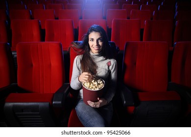 Portrait d'une jolie jeune femme assise dans un théâtre vide, mangeant du pop-corn et souriant, vue de face