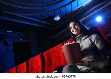 Une jolie jeune femme assise dans une salle de cinéma vide mange du pop-corn en souriant ; derrière elle, on aperçoit le faisceau lumineux bleu du projecteur.