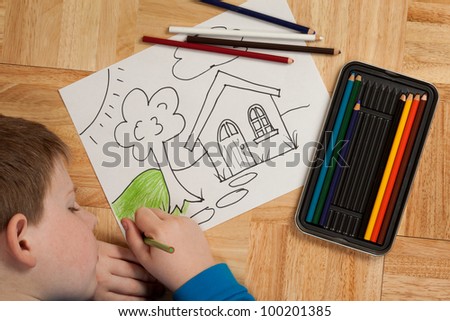 Young boy in blue shirt coloring a picture with pencils while laying on the floor.