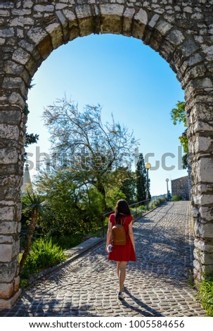 A young woman with a backpack in a red summer dress walking through the ancient, stone arch door. Rijeka, Croatia