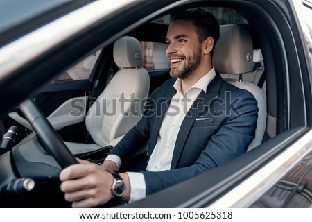 Man of style and status. Handsome young man in full suit smiling while driving a car Royalty-Free Stock Photo #1005625318
