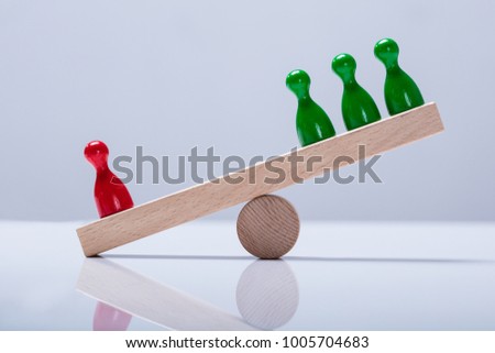 Red And Green Pawns Figures Balancing On Wooden Seesaw Over The Desk