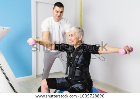 Beautiful senior woman doing exercises in electrical muscular stimulation suit with her personal trainer at rehabilitation center.