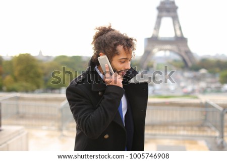 Half Italian and Nigerian man making call with modern smartphone close to Eiffel Tower in slow motion. Young male person has black curly hair, nice smile and earing, wears black coat. Concept of new