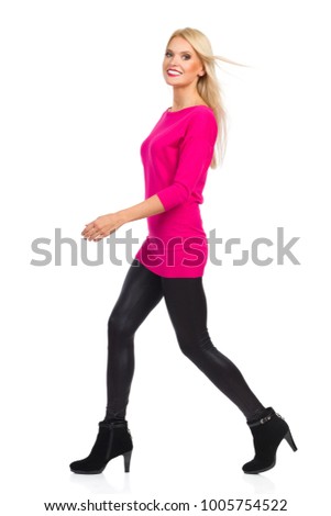 Blond woman in black leggings, high heels and pink sweater is walking, looking at camera and smiling. Side view. Full length studio shot isolated on white.