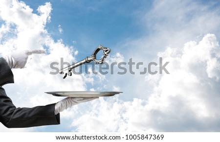 Cropped image of waitress's hand in white glove presenting big key on metal tray and pointing on it with cloudy skyscape on background.