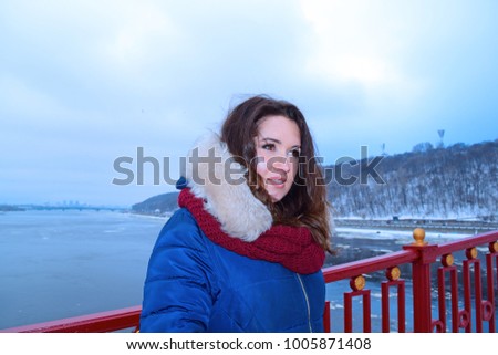 Portrait of young brunette woman in the scarf on the bridge over the river in the city in winter
