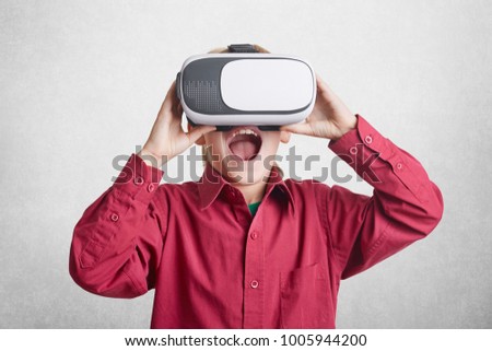 Excited male kid wears virtual relaity goggles, being glad to see fantastic pictures, lives in other world of illusions, isolated over white background. Technology, augmented reality and entertainment