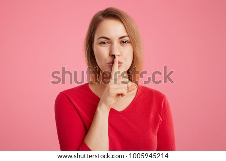 Portrait of secret beautiful young woman keeps fore finger on lips, shows silence sign, tries to keep personal information in secret, isolated over pink background. Hush or stop talking, please