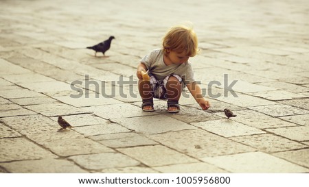 Photo closeup of cute fair-haired blond kid tiny little child baby boy feeding birds with bun sitting on haunches on flag-stone pavement cityscape on blurred grey background, horizontal picture