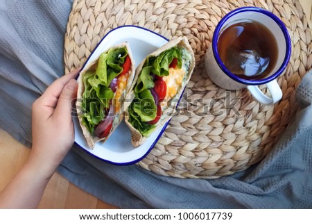 a hand holding a plate of scrambled egg and vegetable pita sandwich on the breakfast table with a mug of black tea