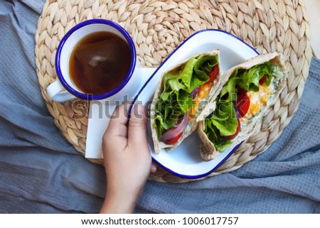 a hand holding a plate of scrambled egg and vegetable pita sandwich on the breakfast table with a mug of black tea